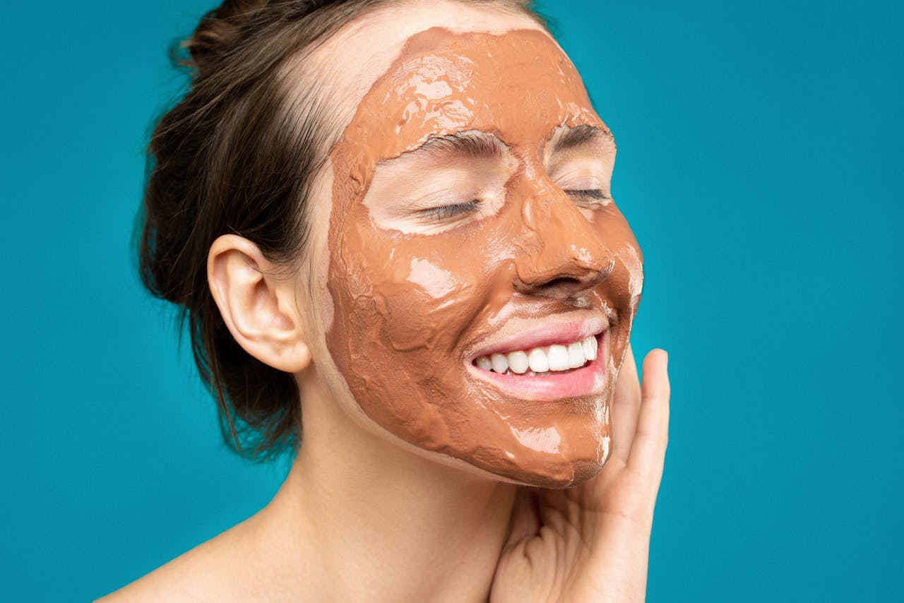 Skincare Confidence | Build Your Glow with Beautiful by Suzie Close-up of a woman smiling with a clay mud mask on her face on a turquoise background.